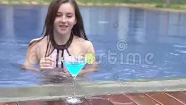 美丽的女孩在游泳池游泳，喝蓝色的鸡尾酒玛格丽塔。